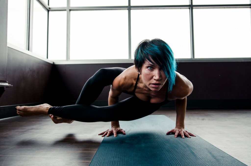 Woman with blue hair performing an advanced yoga pose on a mat indoors, showcasing strength and flexibility.